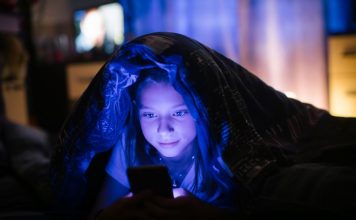 Little girl in bed under a blanket looking at the smartphone at night.